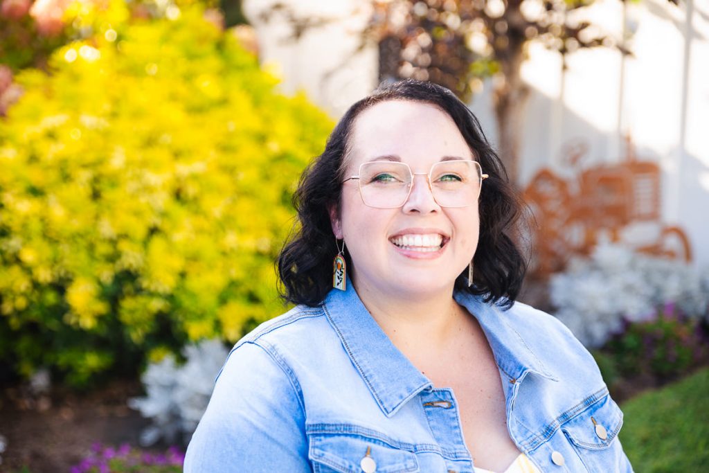 Smiling Caucasian woman wearing a denim jacket and glasses with dark hair, sitting in front of shrubs