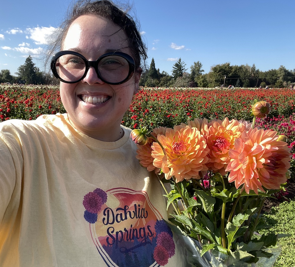 Caucasian woman smiling and wearing black glasses. She's holding a bouquet of orange, pink and yellow dahlias and standing in front of a dahlia field and blue skies.